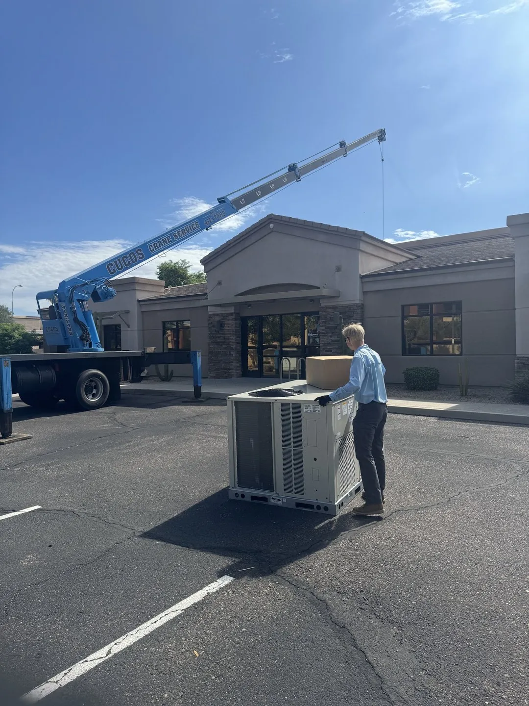A crane lifting a new rooftop package unit onto a commercial building while a Helping Hands technician guides it