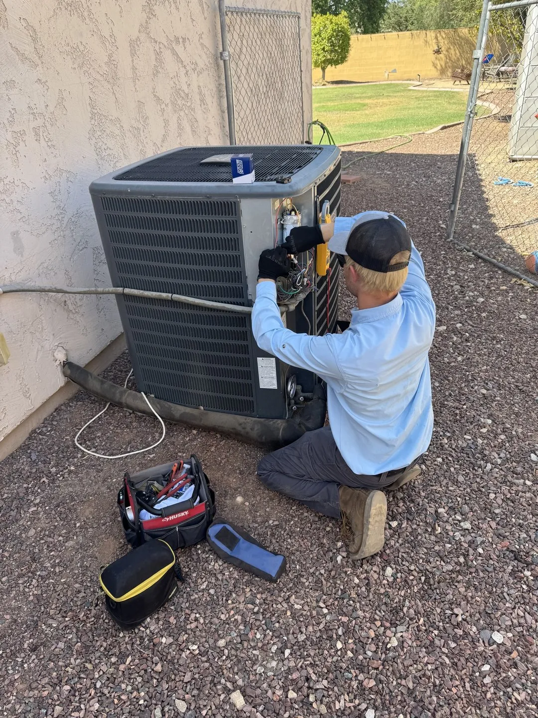 Helping Hands technician kneeling at an outdoor AC condenser with the panel off, testing components